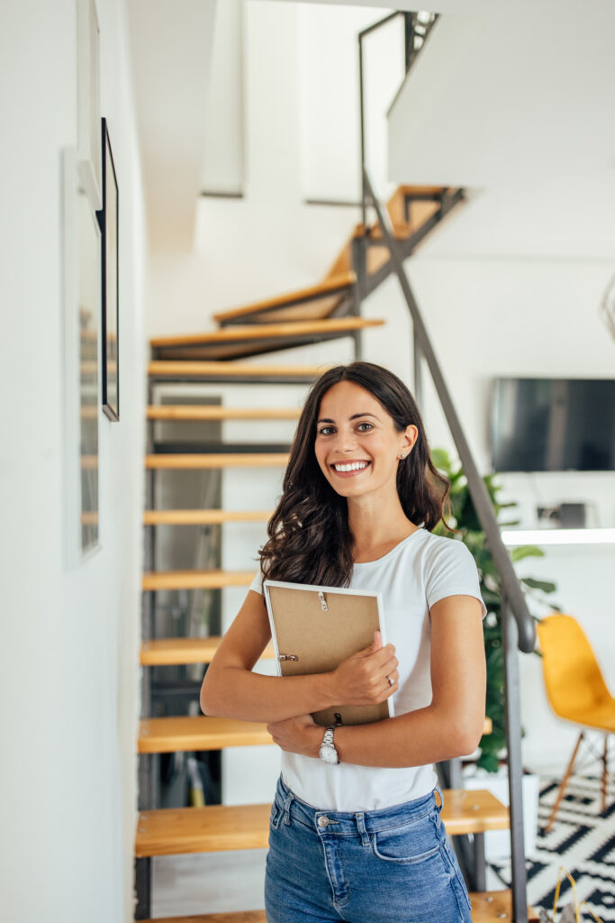 Smiling woman with long dark hair holding a closed cardboard box, standing in a modern, well-lit interior with a wooden staircase and various furnishings in the background. She's wearing a white shirt and blue jeans.