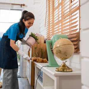 Two people wearing blue shirts and aprons clean a room. The person in the foreground is dusting a shelf with a pair of gloves, while another person in the background carries a black trash bag. A globe and decorative items are on the shelf.