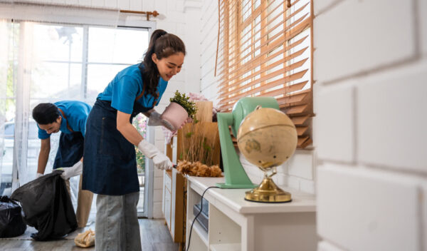 Two people wearing blue shirts and aprons clean a room. The person in the foreground is dusting a shelf with a pair of gloves, while another person in the background carries a black trash bag. A globe and decorative items are on the shelf.