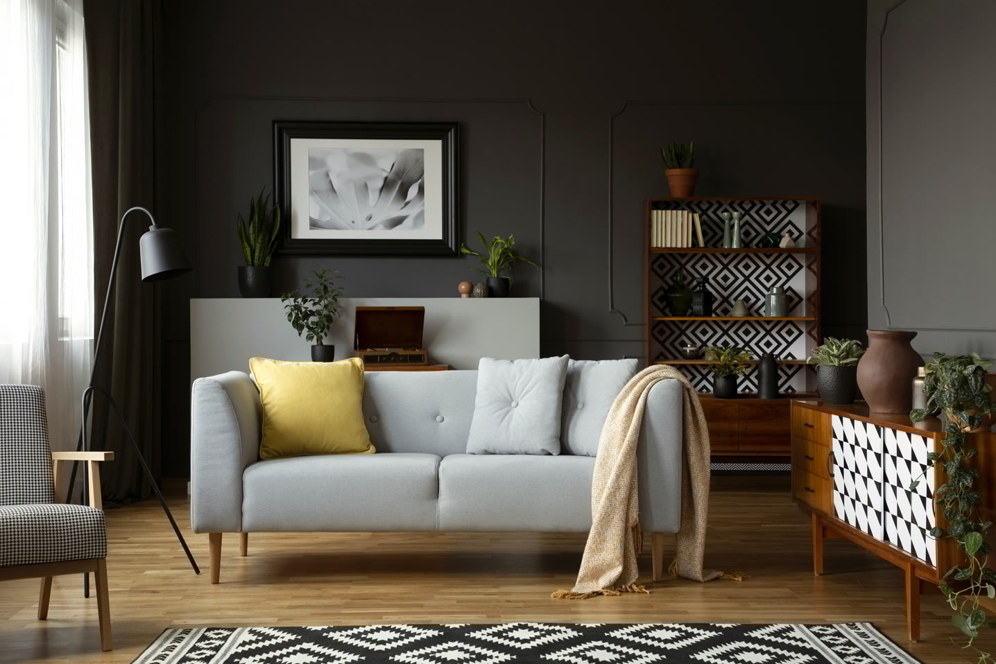 A modern living room with a gray sofa featuring yellow and light gray cushions, a beige throw, and a patterned rug. There are plants, a lamp, a wooden bookshelf, a sideboard, and a black-and-white framed artwork on a dark wall.