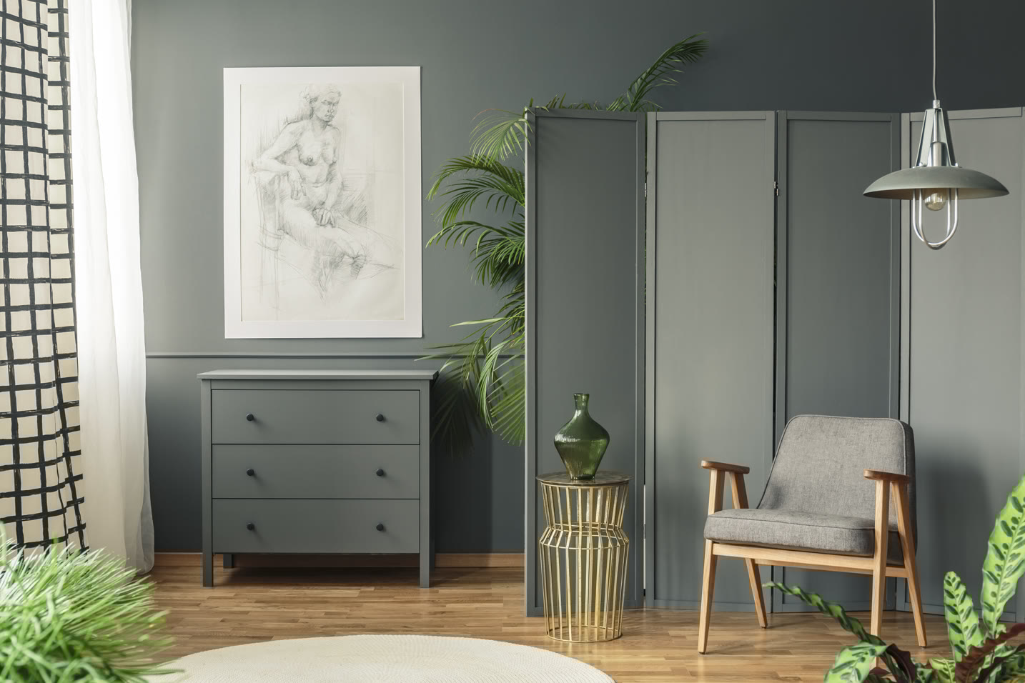 Cozy room with gray walls featuring a dresser and a framed sketch. A chair sits beside a small table with a green vase. A folding screen adds privacy, and plants bring life to the space. Natural light streams through a window with black grid curtains.