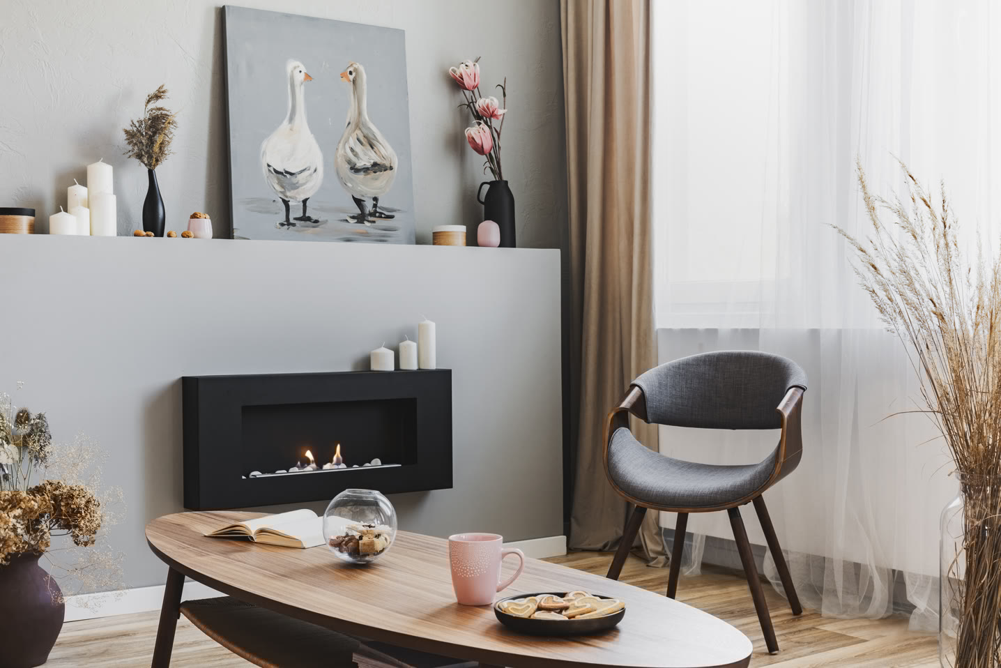 A cozy living room with a modern fireplace on a gray wall. Above it, there is a painting of two geese. A wooden chair sits by a window with sheer curtains. A coffee table with a pink mug, cookies, and a decorative jar is in the foreground.