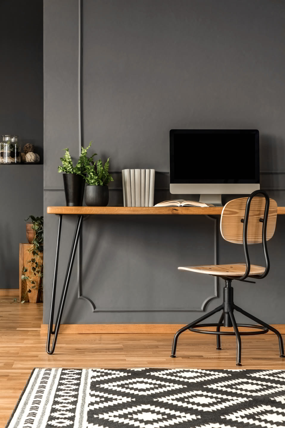 A minimalist home office features a wooden desk with black metal legs, an office chair, a computer, books, and potted plants against a dark gray wall. The wooden floor is partially covered by a black and white geometric-patterned rug.
