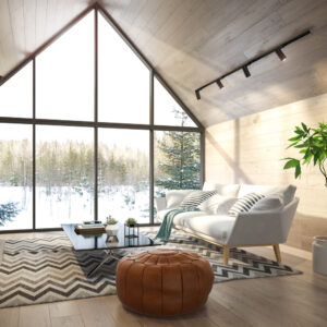 Bright living room with large A-frame windows revealing a snowy forest view. It features a white couch, a modern coffee table, a chevron-patterned rug, a brown leather pouf, and a potted plant. Track lighting lines the ceiling.