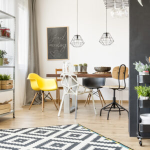 Modern dining room with a mix of chairs around a wooden table, a "good morning" chalkboard, hanging geometric lamps, and a black shelving cart with plants. A metal shelf with plants and storage is on the left. Light wood floor and patterned rug.