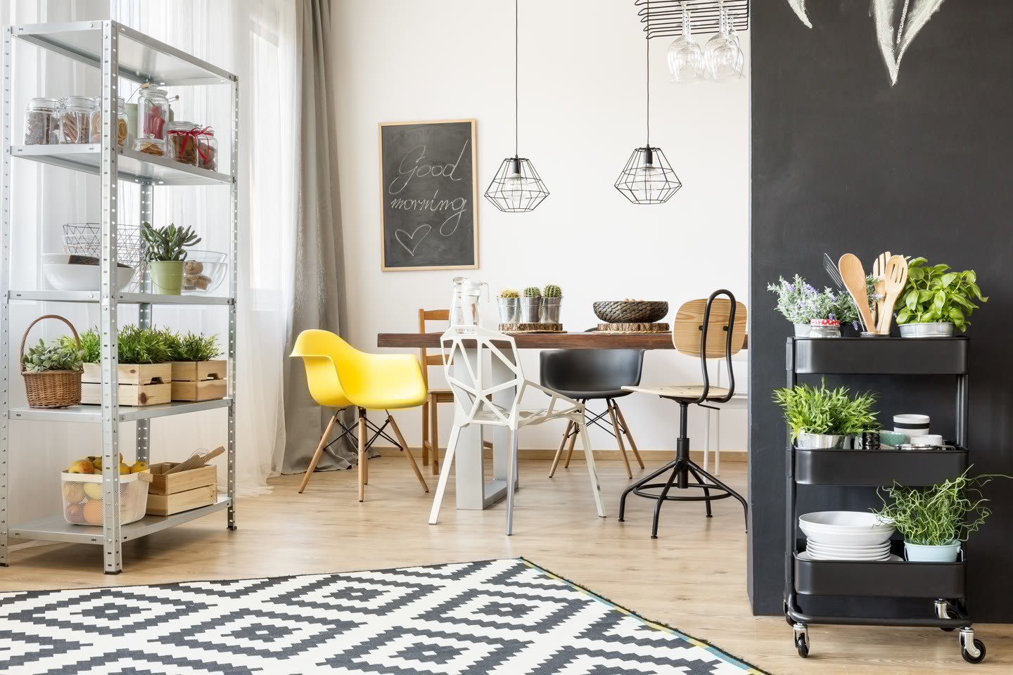 Modern dining room with a mix of chairs around a wooden table, a "good morning" chalkboard, hanging geometric lamps, and a black shelving cart with plants. A metal shelf with plants and storage is on the left. Light wood floor and patterned rug.