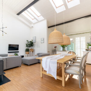 A modern, spacious living room with high ceilings and skylights. It features a wooden dining table with wicker pendant lights, gray sofas, plants, and a wall-mounted TV. The floor is wooden, and there is natural light streaming in.