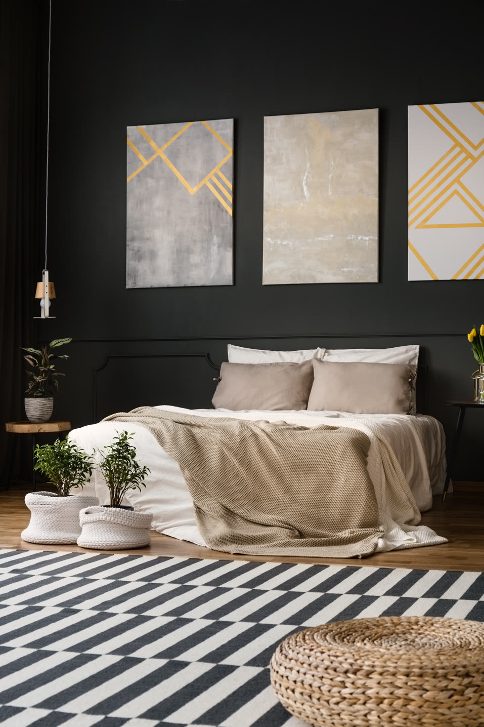 A modern bedroom with a dark wall features a bed with beige pillows and a cream blanket. Above the bed are three abstract paintings with gray and yellow geometric designs. A striped black and white rug and potted plants decorate the floor.
