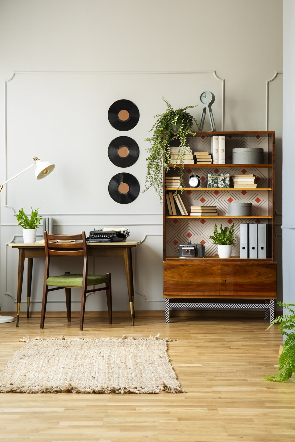 A stylish retro room with a desk and green chair, a vintage typewriter, and a reading lamp. A wooden bookshelf holds books, plants, and a camera. Three vinyl records are hung on the wall. A woven rug lies on the wooden floor.