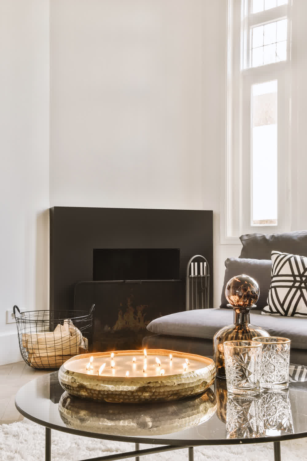 A cozy living room with a modern fireplace and a gray sofa adorned with patterned cushions. A round glass coffee table holds a lit candle centerpiece and decorative glassware. A basket with logs sits nearby on a light rug.