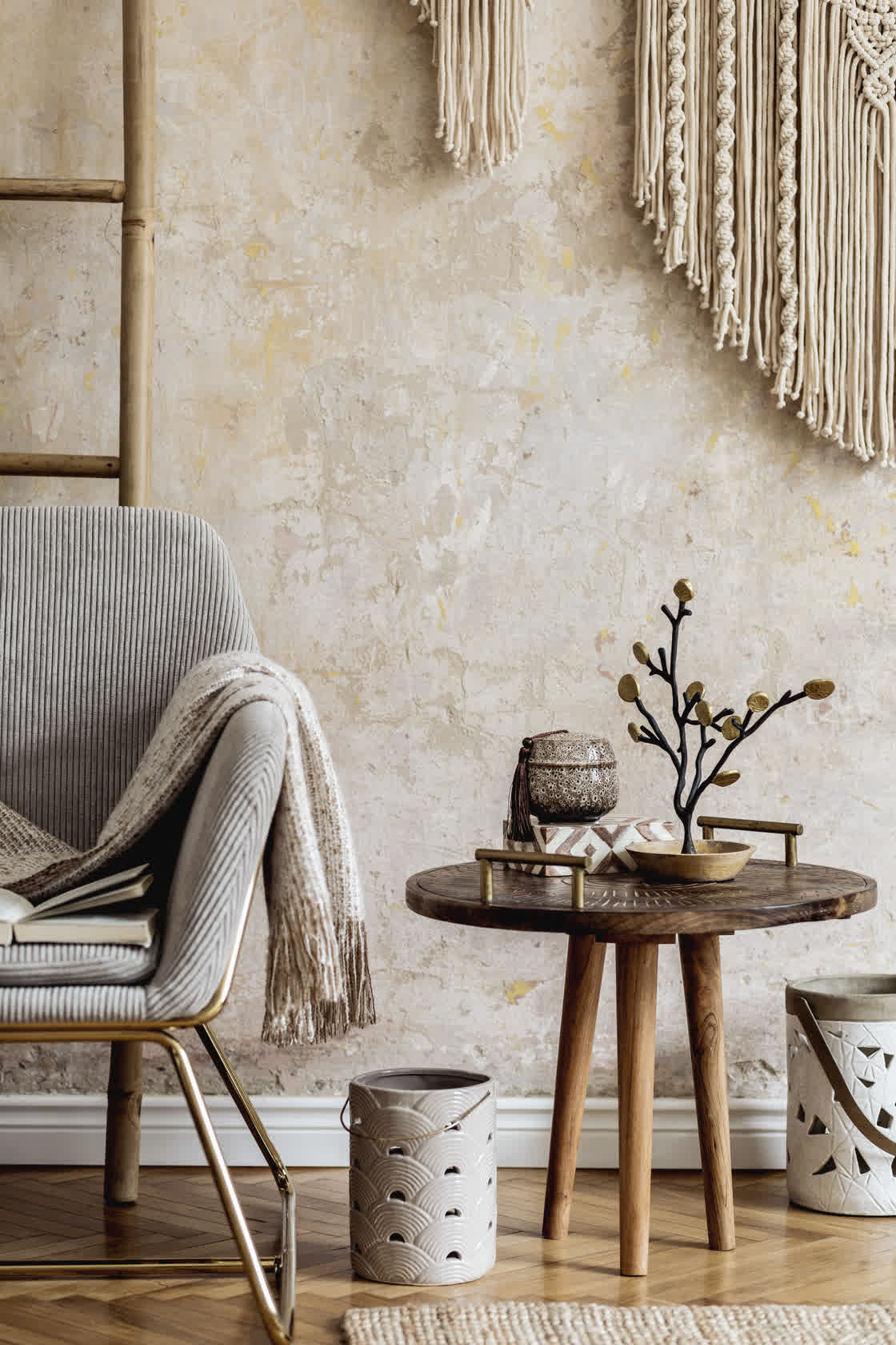 A cozy room with a modern armchair draped with a blanket, a wooden side table holding a decorative branch and candle, and a textured wall with macramé art. A ladder and patterned baskets add to the earthy, minimalist decor.